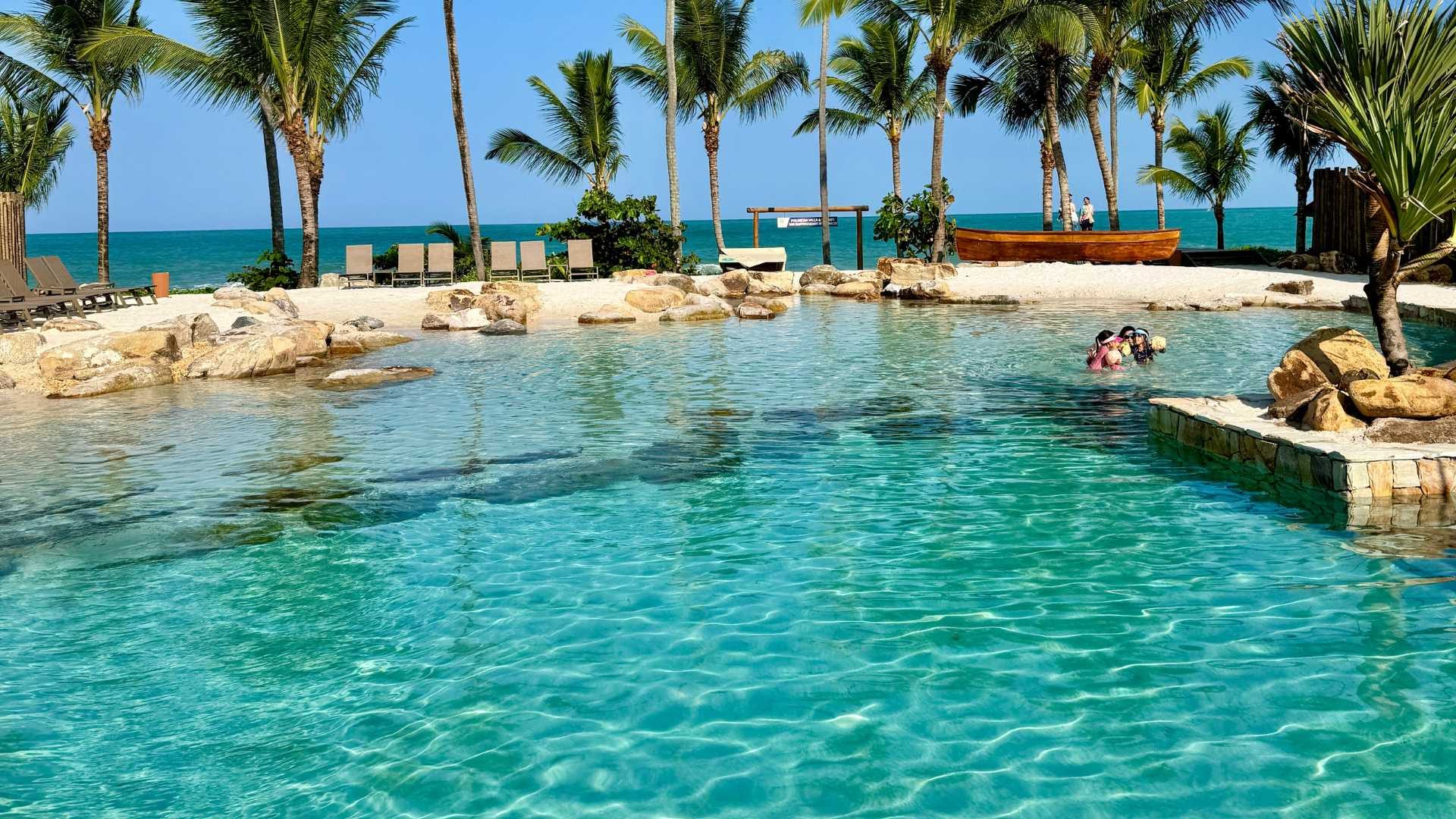 Praia interna Genesis Pool, com água transparente, seixos na borda e o mar ao fundo.