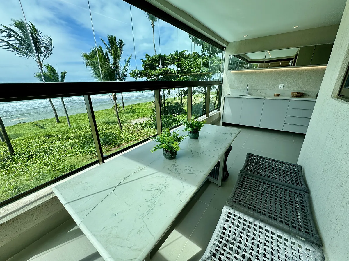 Apartamento La Fleur Collection: varanda gourmet com bancada em m\u00e1rmore e o mar vis\u00edvel atrav\u00e9s do fechamento em vidro.