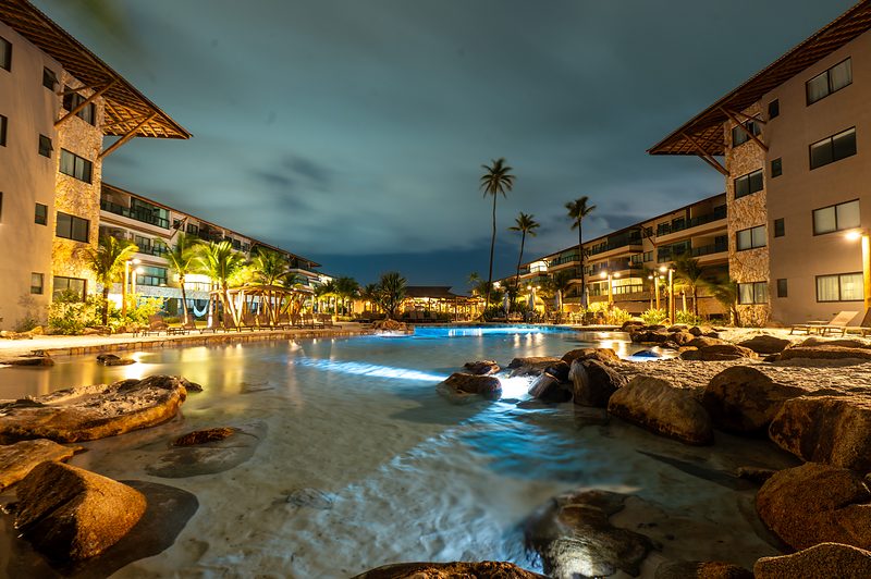 Piscina de borda de areia iluminada \u00e0 noite entre os blocos residenciais do resort.