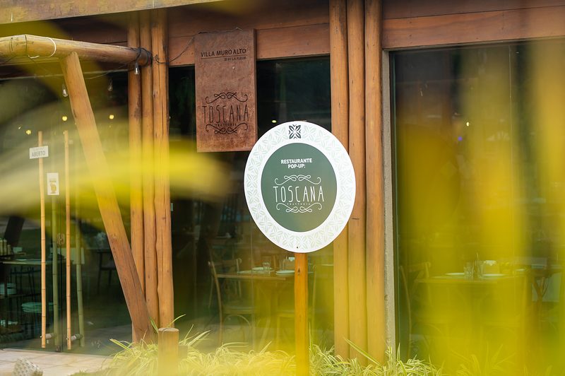 Fachada do restaurante pop-up Toscana na Villa Muro Alto, com placa de madeira e estrutura de bambu.